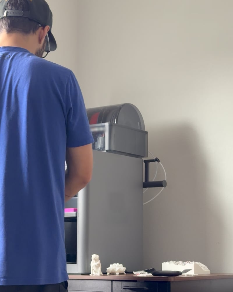 The founder of SideBox Studios, seen from behind in a blue T-shirt and cap, working at a 3D printer with its filament unit on top and small white prints on the cabinet beside it.