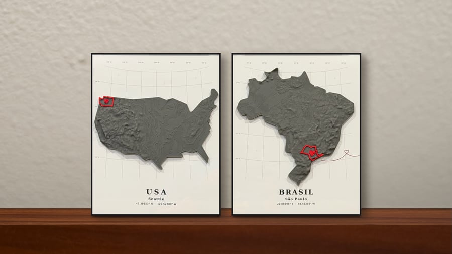 Two black-framed grey relief maps of the USA and Brazil leaning on a wall above a walnut dresser, with Seattle and São Paulo outlined in red and joined by a red heart line.
