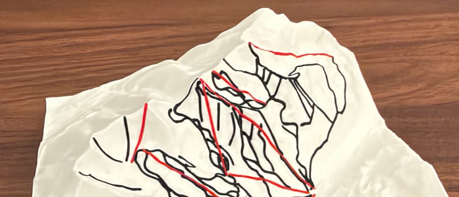 Close view of a white 3D-printed ski mountain relief, its peaks traced with raised black run lines and red lift lines, on a wooden table.