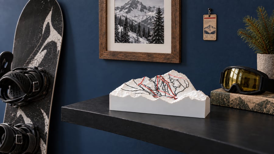 White 3D-printed ski-mountain relief with black and red run lines on a dark shelf, beside a snowboard, a framed mountain print and ski goggles against a navy wall.