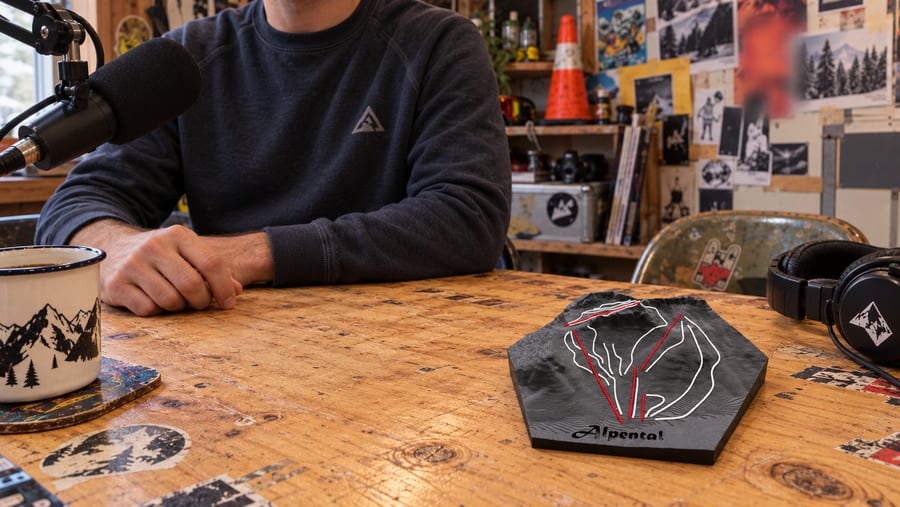 Illustrative scene of a black hexagonal Alpental ski relief with raised white runs and red lift lines on a sticker-covered wooden podcast desk, beside a microphone, an enamel mug and headphones.