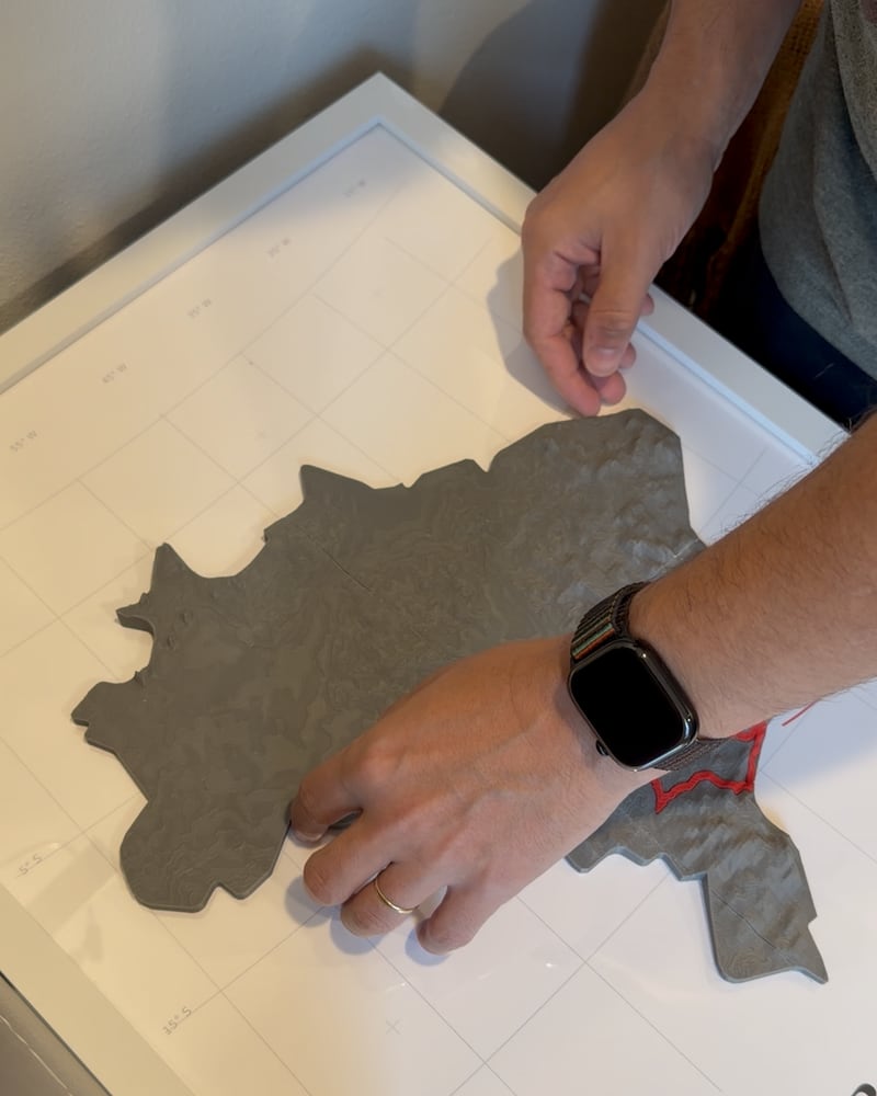 Hands pressing a grey printed Brazil relief onto the map sheet inside a white frame.