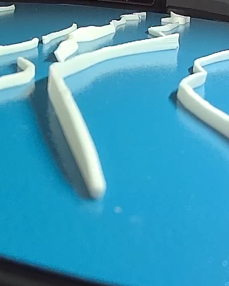 White run-line inlays printing as separate pieces on a blue build plate, one trail standing tall in the foreground.